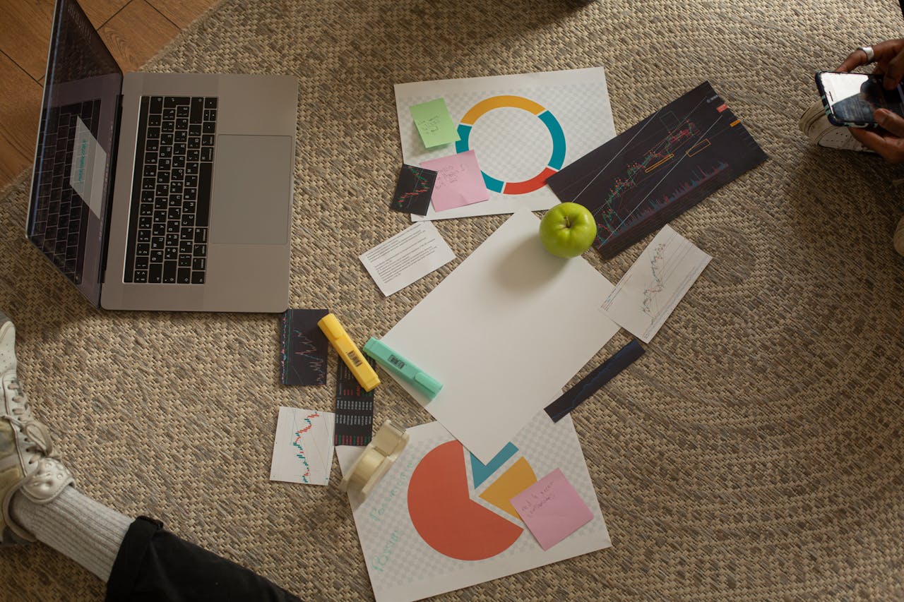 Top view of a workspace with a laptop, charts, sticky notes, and an apple on a woven rug floor.
